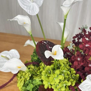 May include: A floral arrangement featuring white calla lilies and anthuriums, vibrant green hydrangea clusters, and deep burgundy flowers. The composition includes dark red foliage and slender green stems, creating a dense, multi-textured display of contrasting colors and botanical shapes.