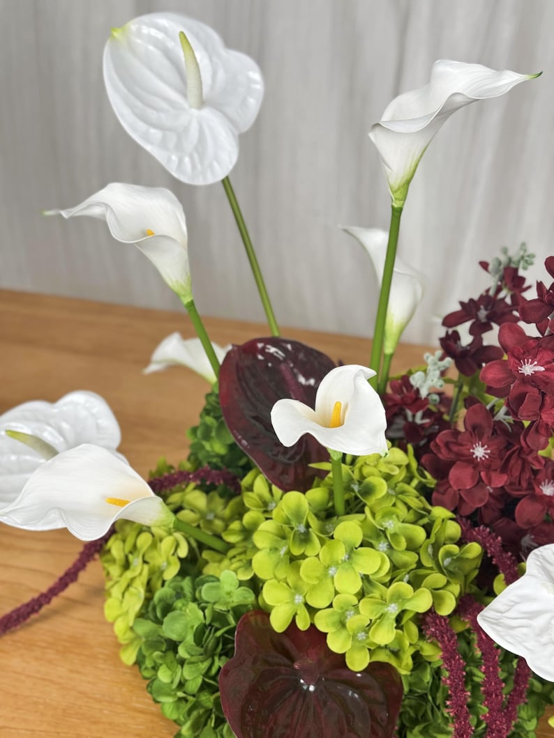 May include: A floral arrangement featuring white calla lilies and anthuriums, vibrant green hydrangea clusters, and deep burgundy flowers. The composition includes dark red foliage and slender green stems, creating a dense, multi-textured display of contrasting colors and botanical shapes.