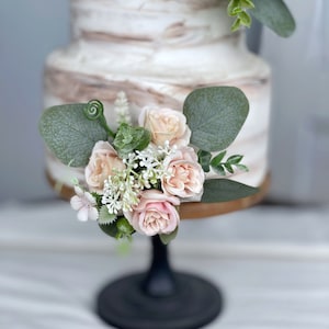 May include: A two-tier wedding cake decorated with a marbled buttercream frosting. The cake is adorned with a cluster of blush pink roses and eucalyptus leaves.