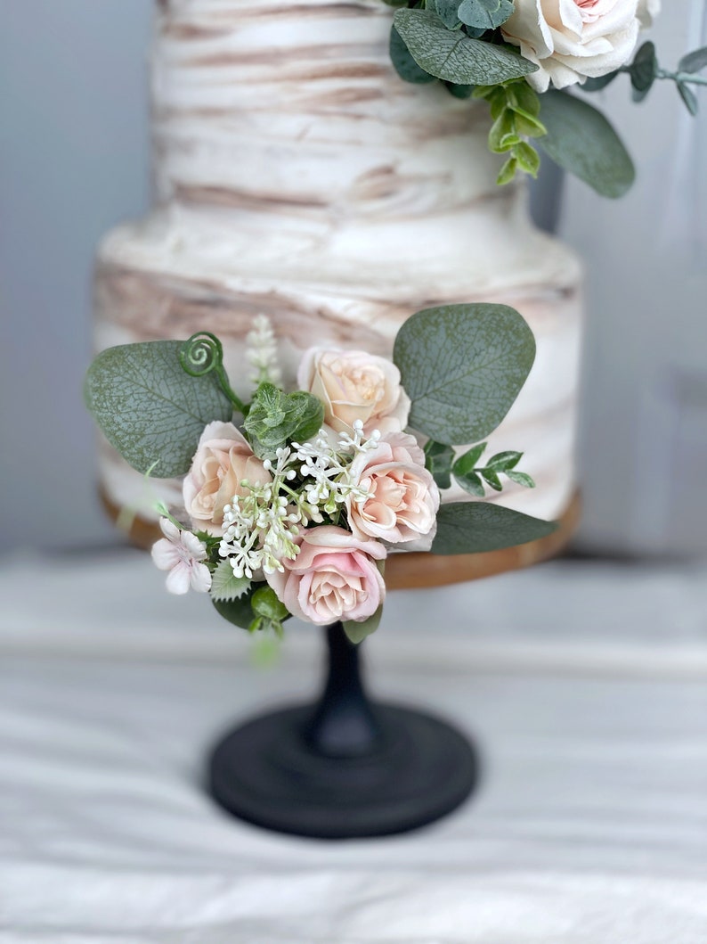 May include: A two-tier wedding cake decorated with a marbled buttercream frosting. The cake is adorned with a cluster of blush pink roses and eucalyptus leaves.