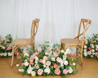 May include: Two wooden chairs with woven seats are arranged in front of a floral display. The flowers are a mix of pink, white, and orange blooms. The background is a white curtain.
