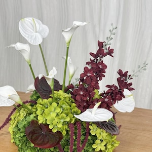 May include: A floral arrangement featuring white calla lilies and white anthuriums, deep burgundy blossoms, and vibrant lime green hydrangea clusters. Long, thin, dark red hanging amaranth stems drape over the base of the bouquet.