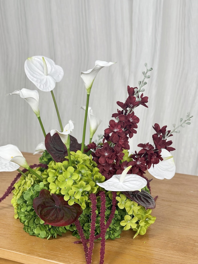 May include: A floral arrangement featuring white calla lilies and white anthuriums, deep burgundy blossoms, and vibrant lime green hydrangea clusters. Long, thin, dark red hanging amaranth stems drape over the base of the bouquet.