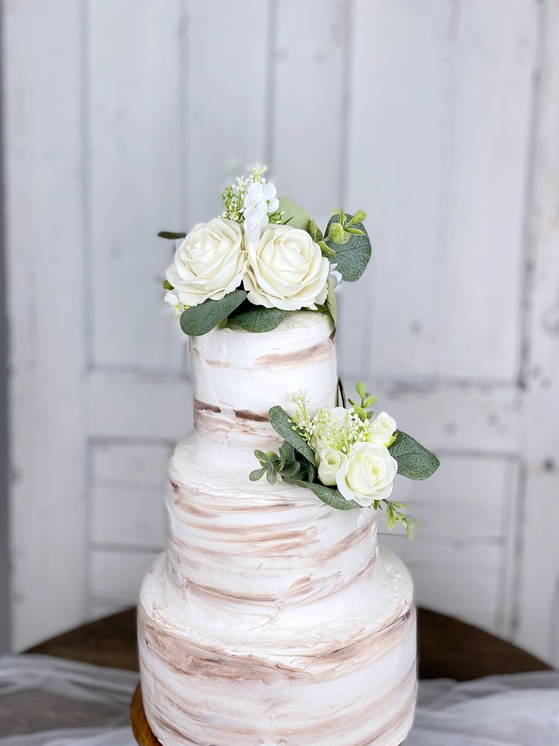 Op de afbeelding: Een drielaagse bruidstaart met witte botercr&egrave;me frosting en bruine marmering. De bovenste laag is versierd met witte rozen en groen.