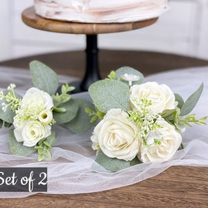 Op de afbeelding: Set van twee witte rozenboeketten met groen, perfect voor bruiloftsdecoratie. De boeketten liggen op een witte tule stof met de tekst "Set of 2" in zwart.
