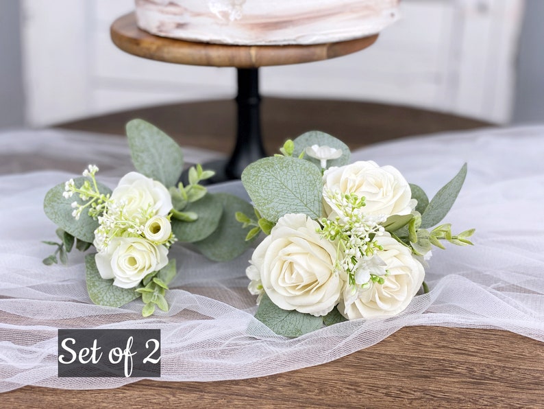 Op de afbeelding: Set van twee witte rozenboeketten met groen, perfect voor bruiloftsdecoratie. De boeketten liggen op een witte tule stof met de tekst "Set of 2" in zwart.