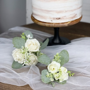 Op de afbeelding: Twee kleine boeketten met witte rozen en groen op een witte tule stof. De boeketten zijn op een houten oppervlak gerangschikt.
