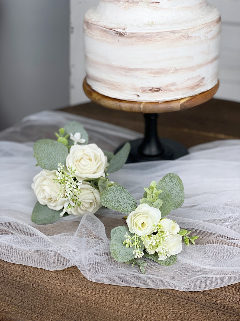 Op de afbeelding: Twee kleine boeketten met witte rozen en groen op een witte tule stof. De boeketten zijn op een houten oppervlak gerangschikt.
