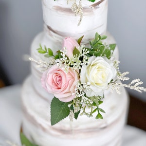 May include: A three-tiered wedding cake decorated with white frosting and subtle brown stripes. Each tier is adorned with a cluster of pink and white roses, greenery, and small white flowers.