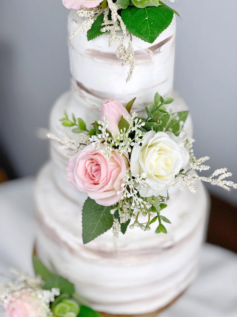 May include: A three-tiered wedding cake decorated with white frosting and subtle brown stripes. Each tier is adorned with a cluster of pink and white roses, greenery, and small white flowers.