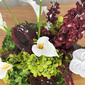 May include: A floral arrangement featuring white calla lilies with yellow centers, deep burgundy anthurium leaves, clusters of small lime green hydrangea-like flowers, and dark red floral sprays with small white centers.