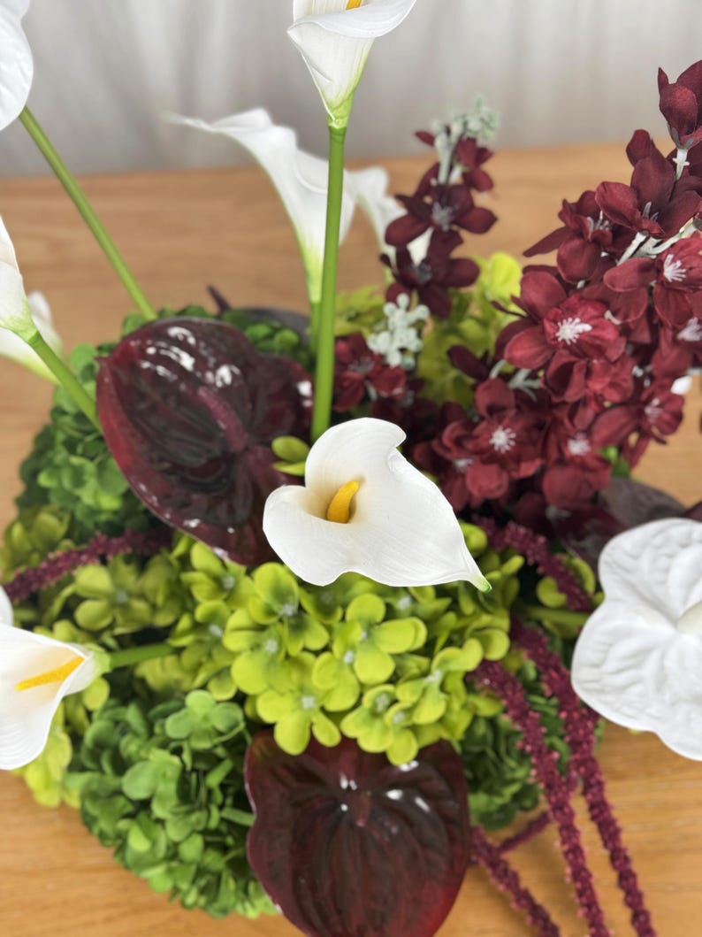 May include: A floral arrangement featuring white calla lilies with yellow centers, deep burgundy anthurium leaves, clusters of small lime green hydrangea-like flowers, and dark red floral sprays with small white centers.