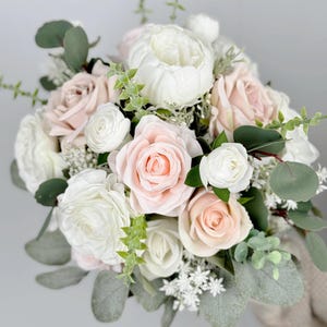 May include: A bridal bouquet featuring white and blush pink roses, white ranunculus, and greenery.