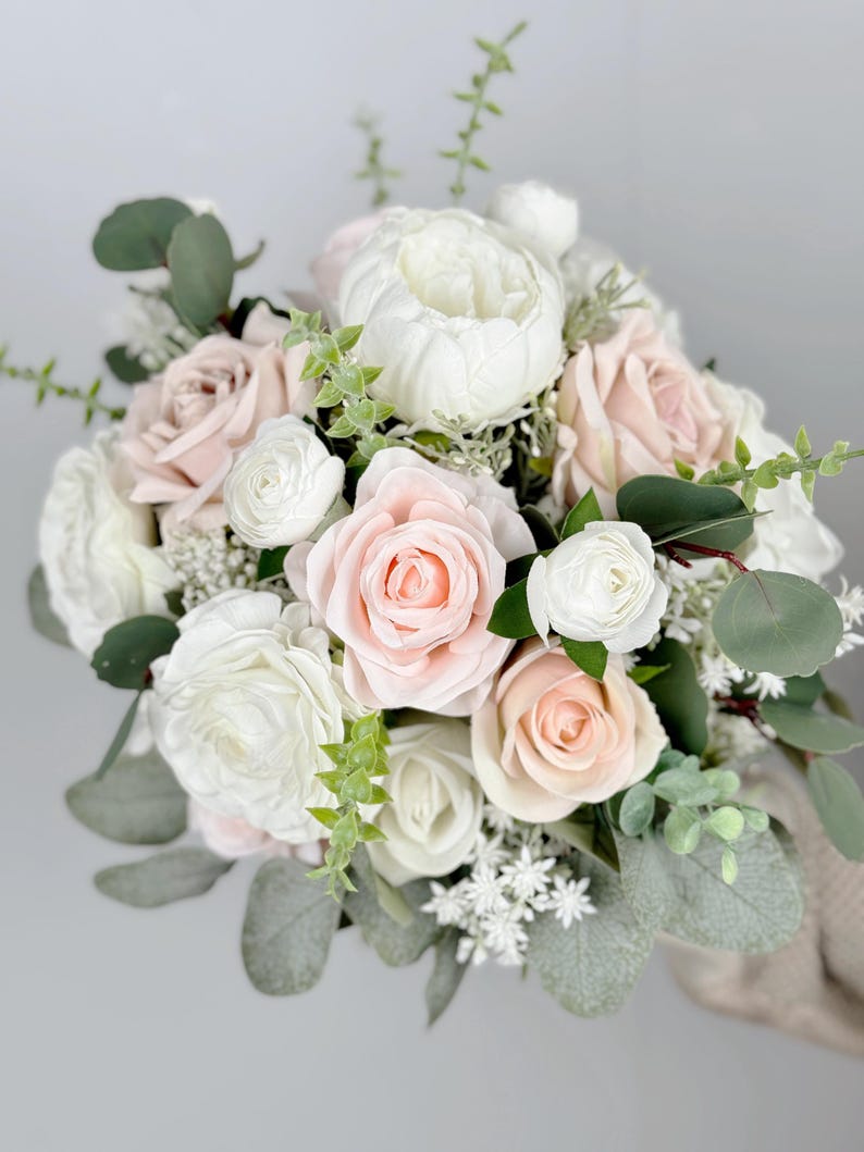 May include: A bridal bouquet featuring white and blush pink roses, white ranunculus, and greenery.