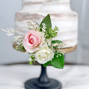 May include: A two-tiered wedding cake with a white and brown marbled frosting. The cake is decorated with a floral arrangement of pink and white roses and greenery. The cake is on a black cake stand.