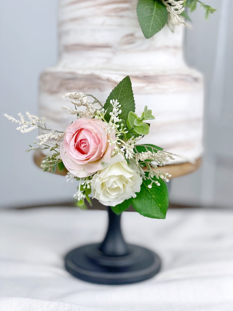 May include: A two-tiered wedding cake with a white and brown marbled frosting. The cake is decorated with a floral arrangement of pink and white roses and greenery. The cake is on a black cake stand.
