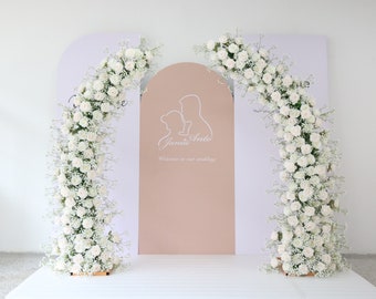 May include: A wedding ceremony backdrop with a white archway decorated with white flowers. The archway is on either side of a pink panel with the text "Jamie & Anto Welcome to our wedding".