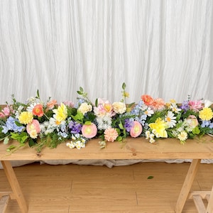 Op de afbeelding: Een houten tafel is versierd met een levendig bloemstuk. De presentatie bevat een verscheidenheid aan kleurrijke bloemen, waaronder roze, gele, oranje en blauwe bloemen, afgewisseld met groen blad. De tafel wordt ondersteund door houten schragen.