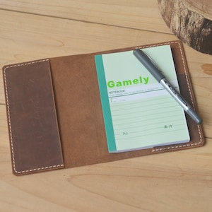 May include: A brown leather notebook cover with a green and white lined notebook inside. The notebook has the word "Gamely" printed on the front and a black pen is lying on top of the notebook.