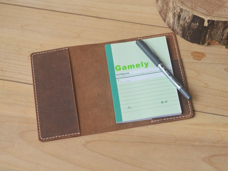 May include: A brown leather notebook cover with a green and white lined notebook inside. The notebook has the word "Gamely" printed on the front and a black pen is lying on top of the notebook.