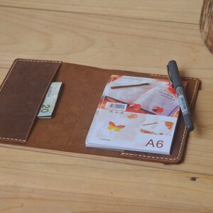 May include: A brown leather notebook cover with a white stitch detail. The cover has a pocket for a pen and a billfold. A small notebook with a butterfly design and the text "A6" is inside the cover.