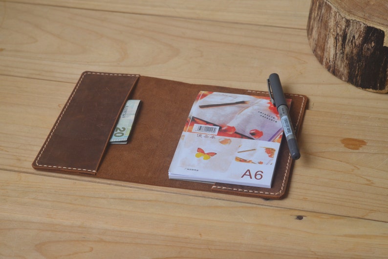 May include: A brown leather notebook cover with a white stitch detail. The cover has a pocket for a pen and a billfold. A small notebook with a butterfly design and the text "A6" is inside the cover.