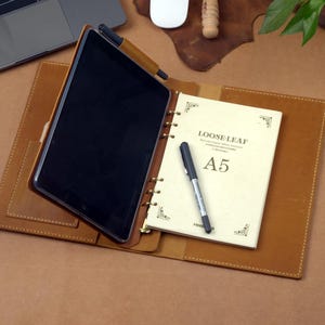 Puede incluir: Un folio de cuero marrón con una tableta y un cuaderno de hojas sueltas. El cuaderno está abierto en una página que dice "LOOSE LEAF" y "A5". Un bolígrafo negro descansa sobre el cuaderno. Un portátil y un ratón son visibles en el fondo.