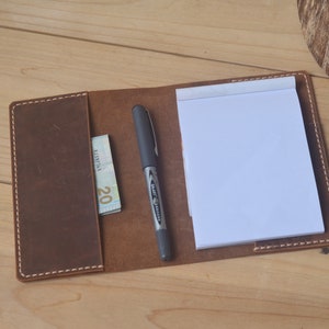 May include: A brown leather notebook cover with a pen and a twenty euro bill inside. The cover has white stitching around the edges.