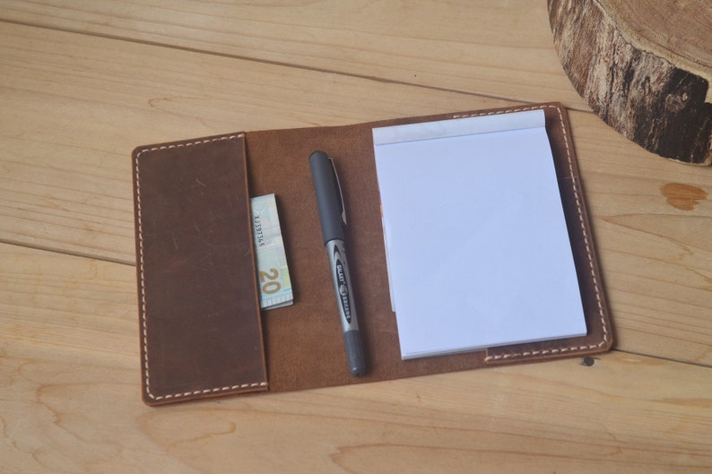 May include: A brown leather notebook cover with a pen and a twenty euro bill inside. The cover has white stitching around the edges.