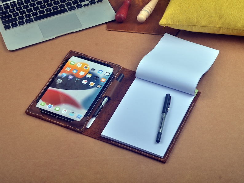 May include: A brown leather portfolio with a tablet, a notebook, and a pen. The tablet is showing a home screen with various apps.