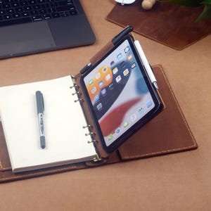 Puede incluir: Un cuaderno y funda para tableta de cuero marrón con un cuaderno blanco, un bolígrafo y una tableta con un lápiz óptico. Un ordenador portátil es visible en el fondo. Los artículos están sobre una superficie marrón.