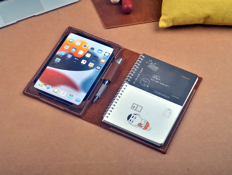 May include: A brown leather folio case with a tablet and a spiral bound notebook. The notebook has a black cover and white pages with handwritten notes and a cartoon drawing.