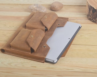 May include: A brown leather laptop sleeve with two front pockets and a flap closure. The sleeve is open and a silver laptop is inside.