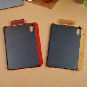 Puede incluir: Dos fundas para teléfono con interior gris oscuro y exterior de cuero de color. Una funda tiene un borde y una pestaña rojos, la otra tiene un borde y una pestaña amarillos. Las fundas son rectangulares con esquinas redondeadas.