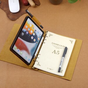 May include: A tan leather-like A5 binder organizer featuring a six-ring metal mechanism, a tablet mounted on the left side, and a loose-leaf paper insert on the right. A black pen is clipped to the binder, and a stylus is held in a side loop.