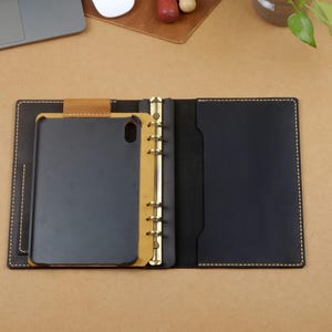 Może przedstawiać: Otwarty sk&oacute;rzany segregator z czarnym etui na tablet i złotymi pierścieniami. Segregator jest otwarty, ukazując czarne wnętrze z kieszenią. Etui na tablet jest czarne i mieści się w segregatorze.
