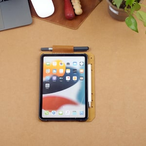 Może przedstawiać: Tablet z czarną ramką i ekranem wyświetlającym ikony aplikacji spoczywa na brązowej sk&oacute;rzanej podkładce. Na podkładce umieszczono rysik i długopis. Widoczny jest r&oacute;wnież laptop, mysz i mała roślina.