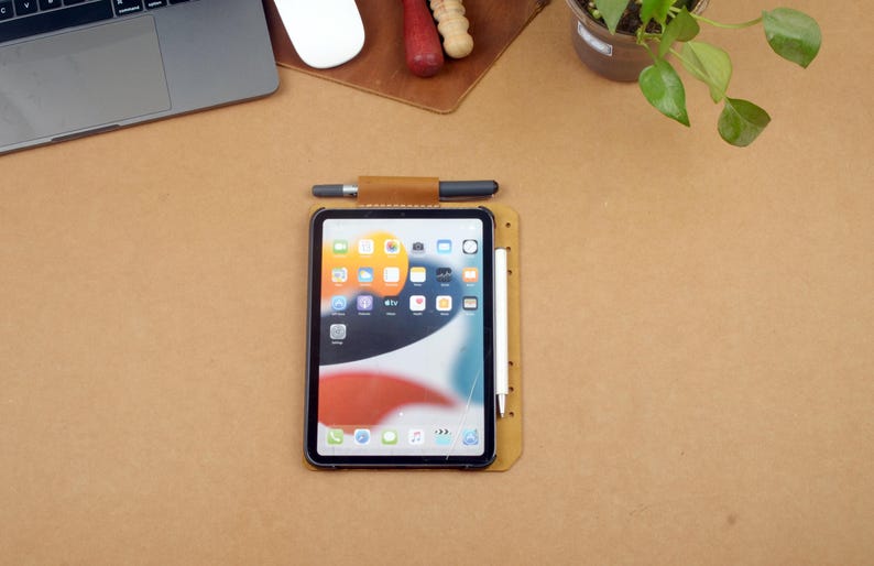 Może przedstawiać: Tablet z czarną ramką i ekranem wyświetlającym ikony aplikacji spoczywa na brązowej sk&oacute;rzanej podkładce. Na podkładce umieszczono rysik i długopis. Widoczny jest r&oacute;wnież laptop, mysz i mała roślina.