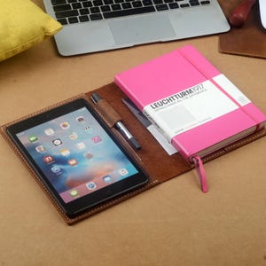 Puede incluir: Una cubierta de cuaderno de cuero marrón con un cuaderno rosa en el interior. La cubierta del cuaderno tiene una ranura para un bolígrafo y una tableta. La cubierta del cuaderno tiene el texto "LEUCHTTURM1917" en la parte delantera.