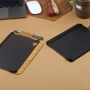 Może przedstawiać: Widoczne są dwa etui na tablet. Jedno etui ma zewnętrzną stronę z jasnobrązowej sk&oacute;ry, czarne wnętrze oraz uchwyt na rysik z umieszczonym w nim rysikiem. Drugie etui jest całkowicie czarne z dopasowaną czarną szlufką na długopis przy g&oacute;rnej krawędzi.