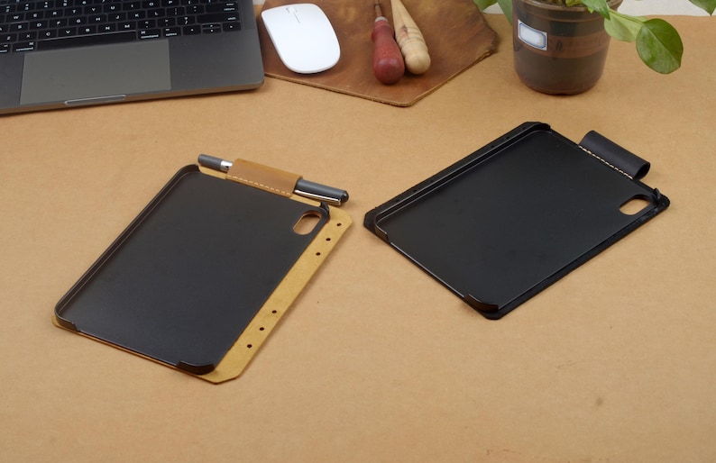 Może przedstawiać: Widoczne są dwa etui na tablet. Jedno etui ma zewnętrzną stronę z jasnobrązowej sk&oacute;ry, czarne wnętrze oraz uchwyt na rysik z umieszczonym w nim rysikiem. Drugie etui jest całkowicie czarne z dopasowaną czarną szlufką na długopis przy g&oacute;rnej krawędzi.