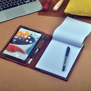 May include: A brown leather portfolio with a tablet, a notebook, and a pen. The tablet is displaying a home screen with various apps.
