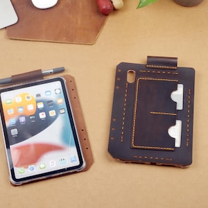 May include: A brown leather tablet case with a pen holder and a separate dark brown leather cover featuring a card slot and yellow stitching. The tablet case holds a tablet with a colourful app icon display, while the cover has two white cards.