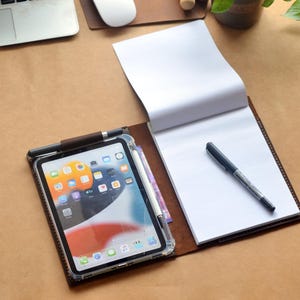 Puede incluir: Un cuaderno de cuero marrón con un bloc de notas blanco y un bolígrafo negro. Un iPad en una funda transparente con un lápiz óptico está al lado del bloc de notas. Un portátil, un ratón y una pequeña planta están en el fondo.
