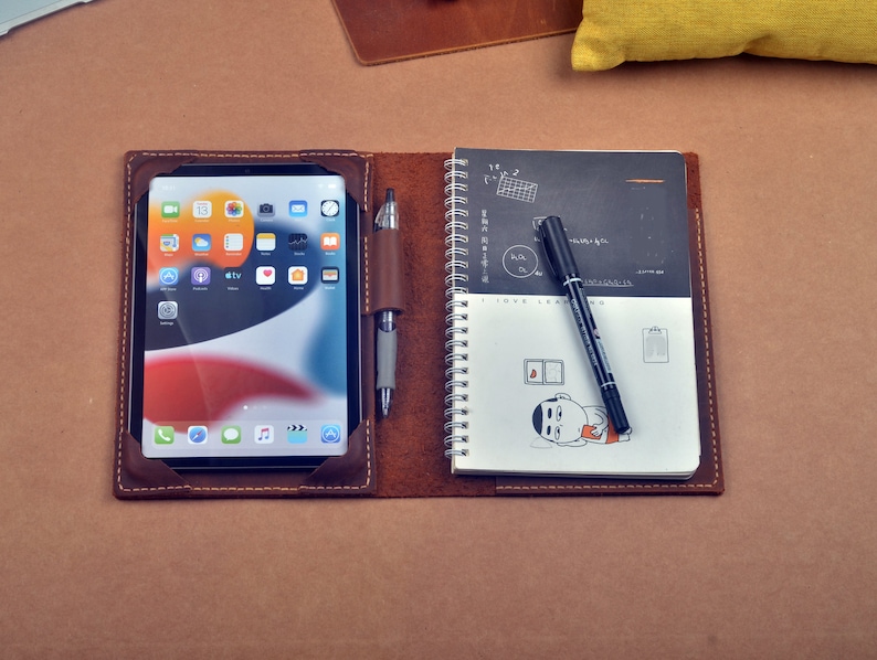 May include: A brown leather notebook with a built-in tablet holder and a black spiral bound notebook. The tablet holder has a black pen and the notebook has a black pen and a doodle of a person.