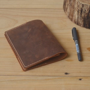 May include: A brown leather notebook with white stitching. The notebook is closed and lying on a wooden surface. A pen is lying next to the notebook.