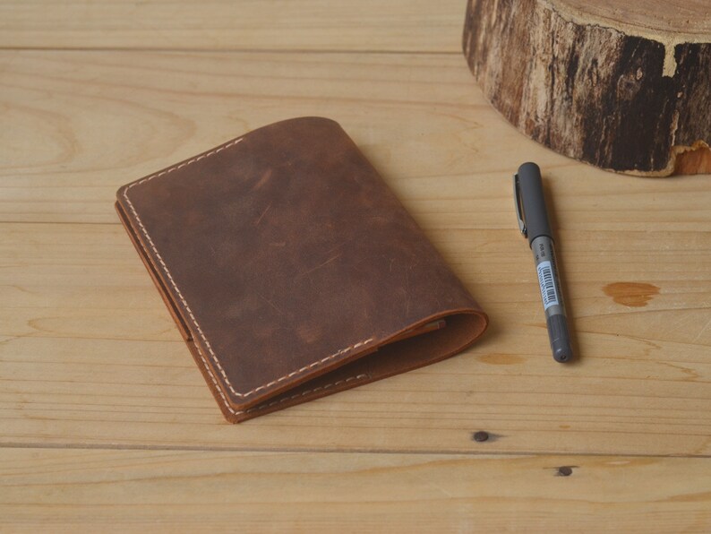 May include: A brown leather notebook with white stitching. The notebook is closed and lying on a wooden surface. A pen is lying next to the notebook.