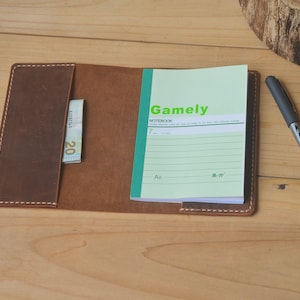 May include: A brown leather notebook cover with a pocket for a bill or card. The cover is open to reveal a lined notebook with the word "Gamely" printed on the front. The notebook is on a wooden surface.