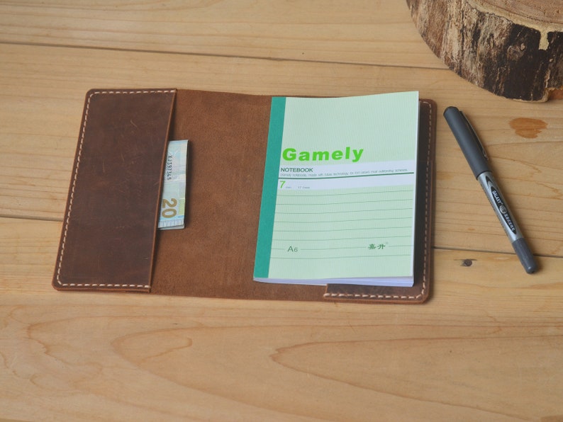 May include: A brown leather notebook cover with a pocket for a bill or card. The cover is open to reveal a lined notebook with the word "Gamely" printed on the front. The notebook is on a wooden surface.