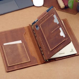Puede incluir: Cuaderno de cuero marrón abierto con múltiples bolsillos y un portalápices. El cuaderno está abierto, revelando un bloc de notas y ranuras para tarjetas. El cuero tiene un aspecto envejecido con costuras visibles.